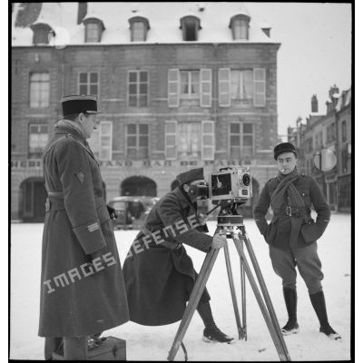 Caméraman du SCA (Service du cinéma aux armées) devant sa caméra sur la place d'un village enneigé.