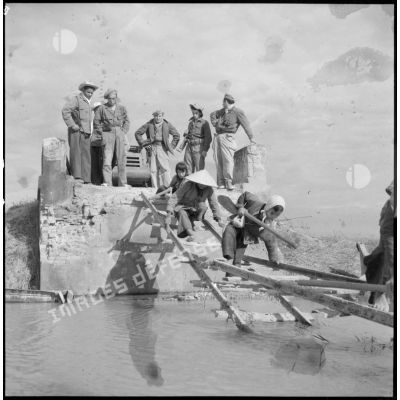 Passage de travailleurs vietnamiens sur des planches de bois remplaçant un pont détruit sur la route coloniale n°1 au Tonkin. A l'arrière-plan, des éléments du Génie.