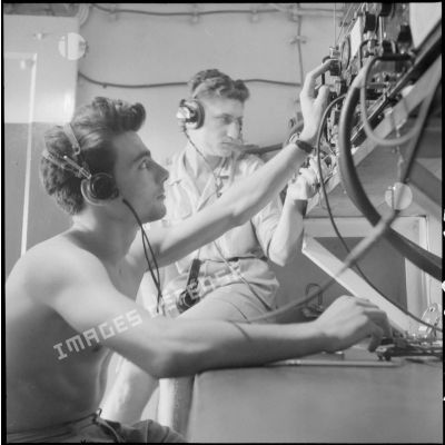 Deux marins, torses nus, au poste de radio pour les opérations de tir.