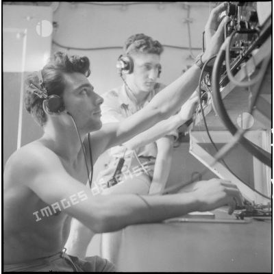 Deux marins, torses nus, au poste de radio pour les opérations de tir.
