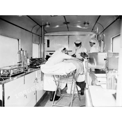 Plan général de l'intérieur d'un camion opératoire, un chirurgien opére secondé par deux infirmières.