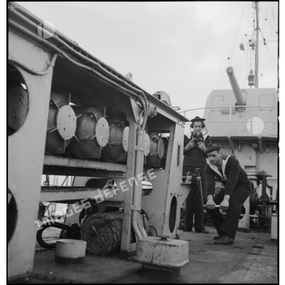 Un matelot manoeuvre le grenadeur de sillage - à chaînes à maillons avec engrenage Galle - de grenades sous-marines à bord du torpilleur léger Branlebas, alors qu'un autre lui communique les ordres de largage.