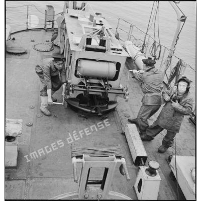 Un second maître et un matelot manoeuvrent le grenadeur de sillage - à chaînes à maillons avec engrenage Galle - de grenades sous-marines à bord du torpilleur léger Branlebas, alors qu'un autre leur communique les ordres de largage.