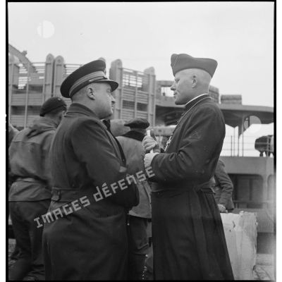Un lieutenant de vaisseau et un aumônier de la marine nationale discutent sur un quai du port de Brest alors que des chasseurs alpins embarquent à bord de navires à destination de l'Ecosse puis de la Norvège.