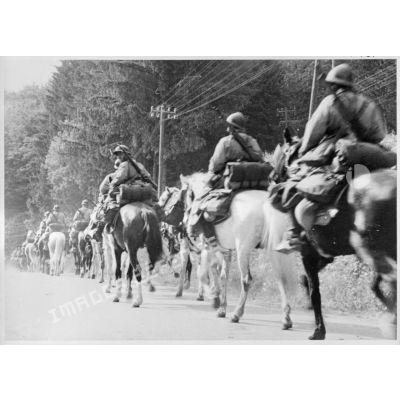 Une colonne de cavaliers à cheval est photographiée le long d'une route de trois quarts dos en plan général.