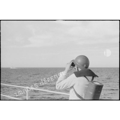 Un marin de quart assure une veille aux jumelles sur le pont du croiseur la Marseillaise. Il porte sur le dos un masque à gaz AFM 34.