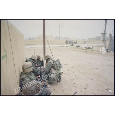 Des marsouins du 3e RIMa (régiment d'infanterie de marine) sont postés accroupis derrière un mur dans une rue d'Al Salman. Deux d'entre eux portent un émetteur-récepteur TRPP 13 au dos.