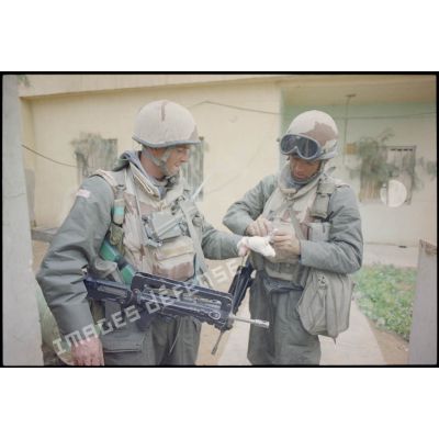Près d'une maison d'Al Salman, un marsouin du 3e RIMa (régiment d'infanterie de marine) soigne un camarade légèrement blessé à la main, qui fume et porte en collier un émetteur-récepteur TRPP-11. Les deux militaires portent leur équipement individuel : casque F1, brelage, gilet pare-éclats ainsi que leur fusil d'assaut FAMAS et le S-3P (survêtement de protection à port permanent) NBC (risque nucléaire, bactériologique et chimique).