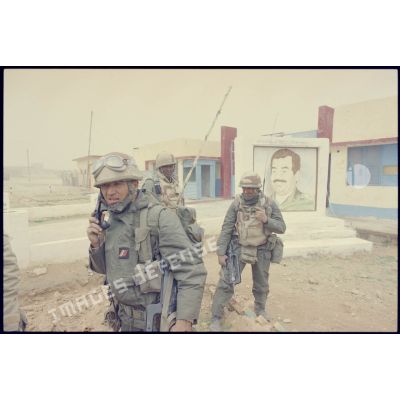 Deux radios du 3e RIMa (régiment d'infanterie de marine), émetteur-récepteur TRPP-13 au dos, se tiennent devant un portrait de Saddam Hussein à Al Salman, alors que deux officiers, dont un lieutenant se concertent. Les marsouins portent leur équipement individuel, un S-3P (survêtement de protection à port permanent) NBC (risque nucléaire, bactériologique et chimique) et leur fusil d'assaut FAMAS.
