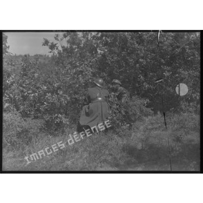 Deux soldats se tiennent postés derrière la végétation, ils sont photographiés de trois quarts dos en plan général.