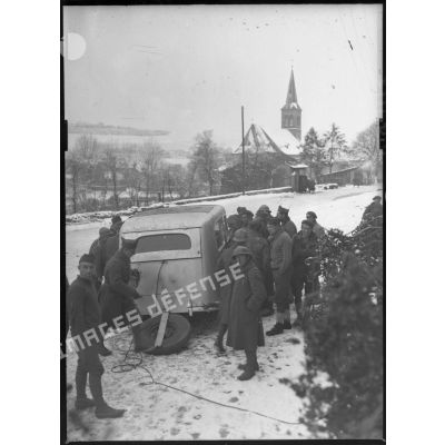 Sous la neige, plan général de soldats qui entourent un véhicule à l'entrée d'un village.