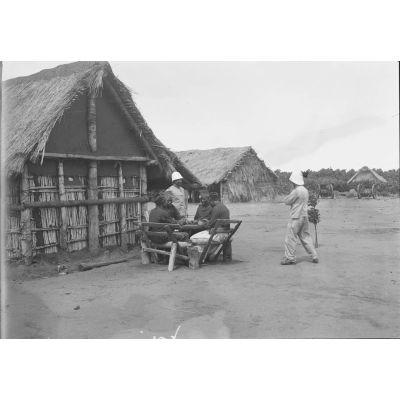 [Des officiers et soldats de l'infanterie coloniale assis à une table dans un village.]