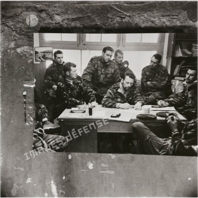 [Photographie de la guerre d'Algérie (1954-1962) scotchée sur un mur, représentant des militaires français rassemblés autour d'un bureau.]