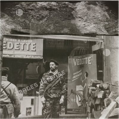 [Photographie représentant un parachutiste devant une affiche publicitaire pour les lave-linges Vedette à Alger, 1954-1962.]