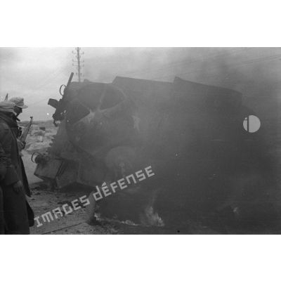 Un char Stuart M3, renversé, brûle au bord d'une route, des soldats allemands s'arrêtent pour l'examiner.