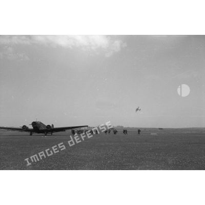 Des troupes s'apprètent à monter dans un avion de transport Junkers Ju-52, en arrière plan dans le ciel de l'aérodrome, un chasseur lourd Messerschmitt Bf-110.