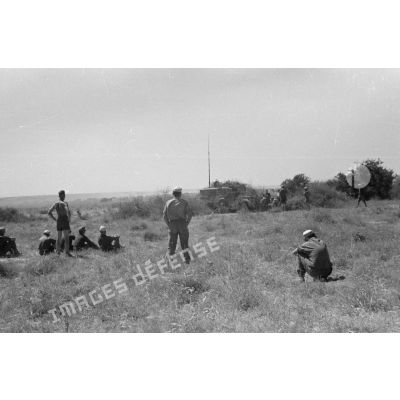 Des soldats assis à même la terre, en demi-cercle, écoutent un cadre torse nu. Tentes et véhicules divers.