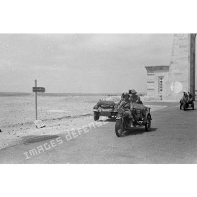 Une voiture allemande Kübelwagen et des mototricycles italiens d'une unité de Bersaglieri passent sous l'Arco dei Fileni sur la via Balbia.