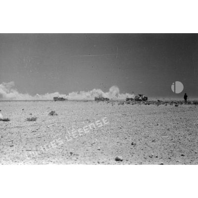 La colonne de véhicules face à un nuage de poussière soulevé par ce qui semble être les explosions d'un tir d'artillerie ou par un vent de sable.