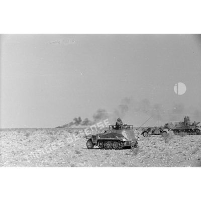 Un semi-chenillé Sd.Kfz-250/3 de l'Art.Rgt-33, une voiture Kfz-16. Un char Panzer IV (Pz-IV) du Panzer-Regiment 8 (Pz.Rgt-8) suivi d'un camion, au loin un véhicule en feu.