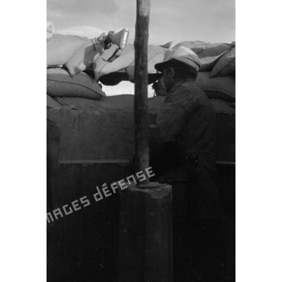 Un soldat observe le terrain depuis une tranchée,. des fusils Mauser (KAR-98k) sont posés sur les sacs de sable du parapet.