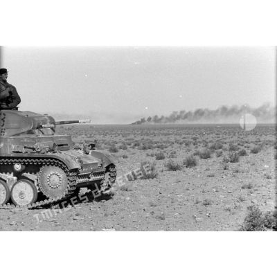 Un char Panzer II (Pz-II) du Panzer-Regiment 8 (Pz.Rgt-8) dans le désert, au loin un nuage de fumée dégagé par un véhicule en flamme.