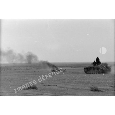Un blindé allemand Panzer III (Pz-III) passe près d'un véhicule en flamme.