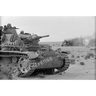 Un Panzer III (Pz-III) passe près d'un camion en flamme.