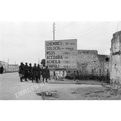 Un groupe de soldats italiens en dessous d'un grand panneau kilomètrique indiquant Msus, Agedabia, ElL-Agheila, Tripoli.