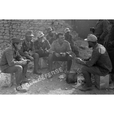 Un groupe de soldats parle et se restaure, près d'un mur en partie éboulé.