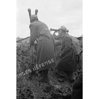 Protégés derrière un parapet ou dans une tranchée, des officiers et un soldat observent les combats avec un binoculaire et un télémètre.
