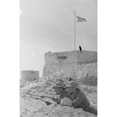 Deux Bersaglieri allongés dans le sable, le fusil au point, en arrière-plan une des redoutes du fort surmontée du drapeau italien.