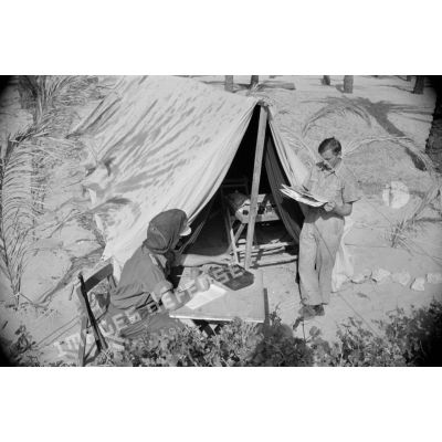 Un Leutnant écrit sur une table, il a recouvert sa casquette d'une moustiquaire, un autre lit près d'une tente.