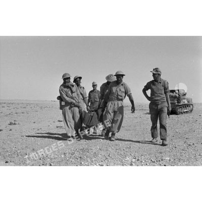 Des prisonniers transportent un blessé installé dans une couverture, gardés par deux soldats allemands armés de pistolets Walther-P-38.