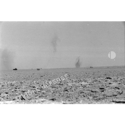 Des véhicules disséminés sur le terrain, dans le lointain des impacts de tir d'artillerie.