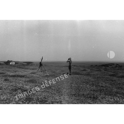 Un soldat observe le terrain à la jumelle près d'une mitrailleuse MG-34 sur trépied.