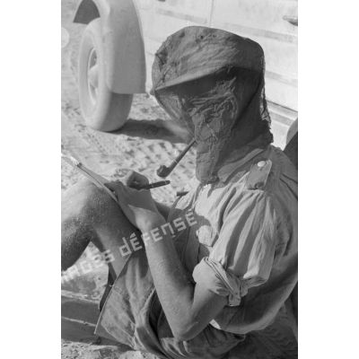 Un soldat écrit en fumant la pipe, la tête protégée par un filet anti-moustique.