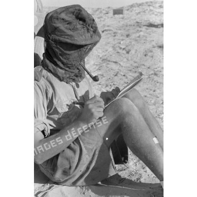 Un soldat écrit en fumant la pipe, la tête protégée par un filet anti-moustique.