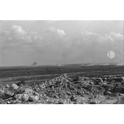 Vue générale du désert. Une colonne blindée progresse avec dans le lointain des impacts de tir d'artillerie.