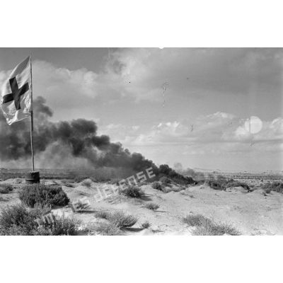 Des débris d'un avion en feu près d'un drapeau de la croix rouge dans le désert.