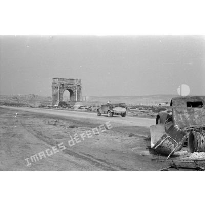 Une voiture Kübelwagen progressant le long de la route, on reconnaît l'Arc de Dioclétien près d'une épave de camion et des ruines romaine de Sufetula.