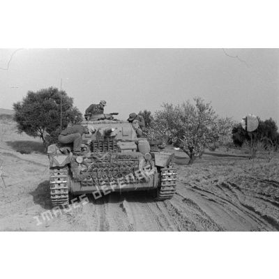 Un char Panzer III (Pz-III Ausf-J) roule sur une piste, une partie de son équipage se tient à l'extérieur de l'engin.