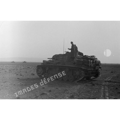 Un char de commandement Panzerbefehlswagen III (PzBefWg-III) roule dans la plaine, dans le lointain, deux chars Panzer IV (Pz-IV).