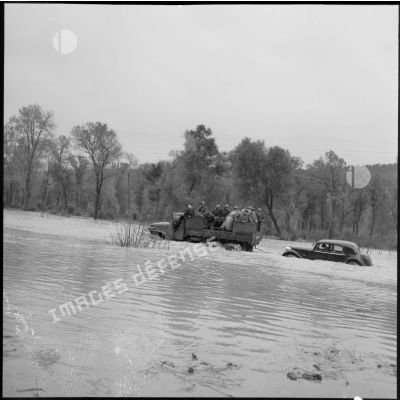 Remorquage d'une Citroën traction, sur un chemin inondé, par une jeep de l'armée avec à son bord des soldats et des officiers supérieurs.