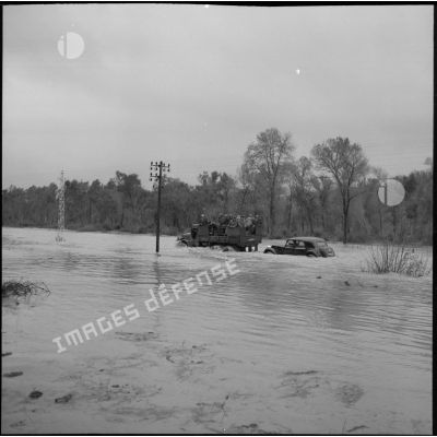 Une jeep de l'armée, avec à son bord des officiers généraux et des soldats, remorque une Citroën traction sur un chemin inondé.