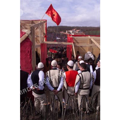 Vue des coulisses avec un groupe folklorique de dos sous un drapeau de l'armée de libération du Kosovo (UCK).