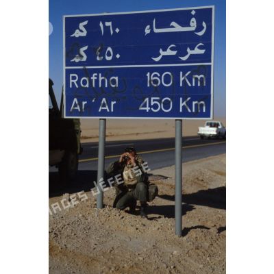 Un soldat accroupi sous un panneau indicateur indiquant la direction de Rafha, observe les abords de la route, sur la Tapline Road (trans-arabian pipeline), axe routier reliant Miramar à la ZDO (zone de déploiement opérationnel) Olive.