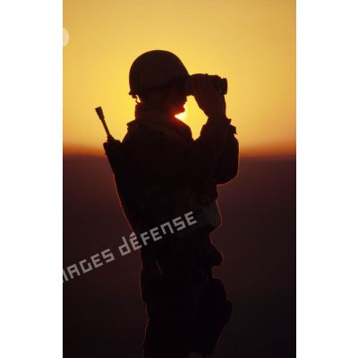 En ZDO (zone de déploiement opérationnel) Olive, un soldat est posté en observation dans le désert, équipé de jumelles, en contre-jour au coucher de soleil.