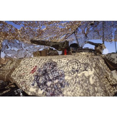 En ZDO (zone de déploiement opérationnel) Olive, tourelle d'un char de combat Abrams-M1 américain, décorée d'un dessin représentant la mort tenant une hache et un loup sur une falaise au clair de lune, sous un filet de camouflage.