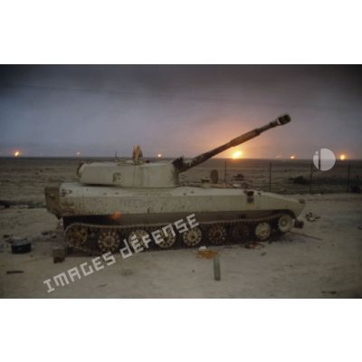 Un canon automoteur irakien de 122 mm M1974 (ou 2S1) abandonné devant un horizon de puits de pétrole en feu.